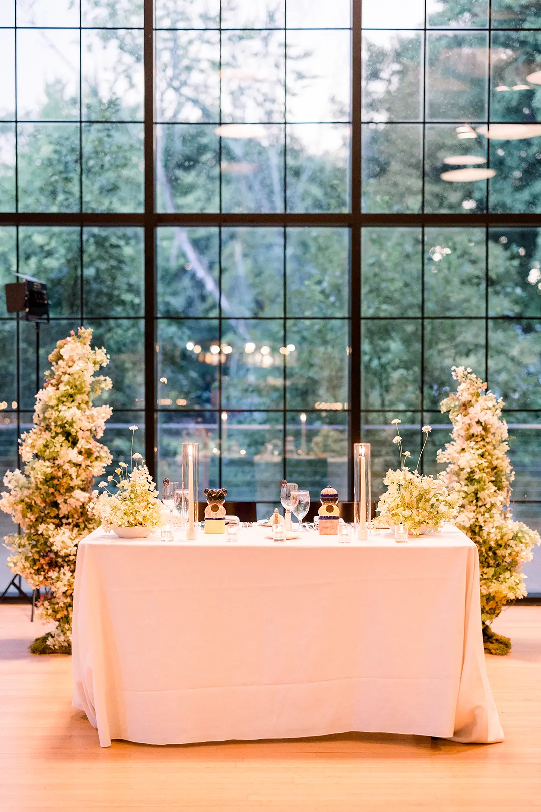 Elegant sweetheart table with white linens, candles, and floral arrangements by large windows.