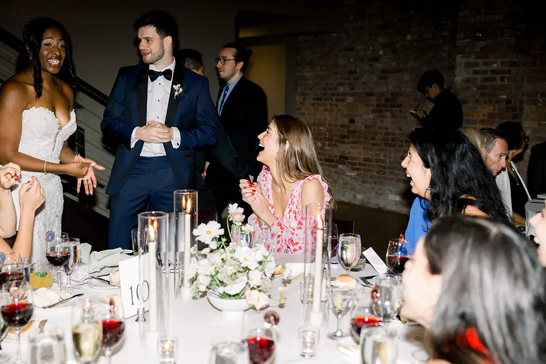 Bride and groom laugh with guests at a candlelit wedding reception table indoors.