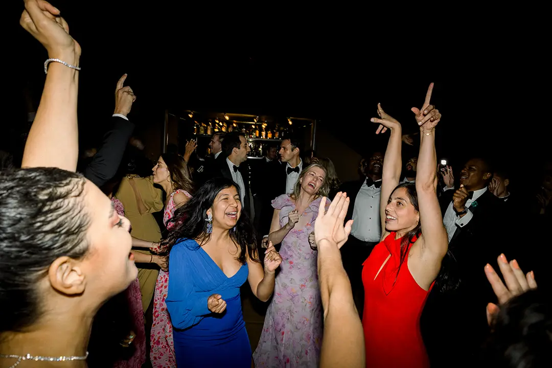 Wedding guests dancing joyfully with raised hands during a lively nighttime reception.
