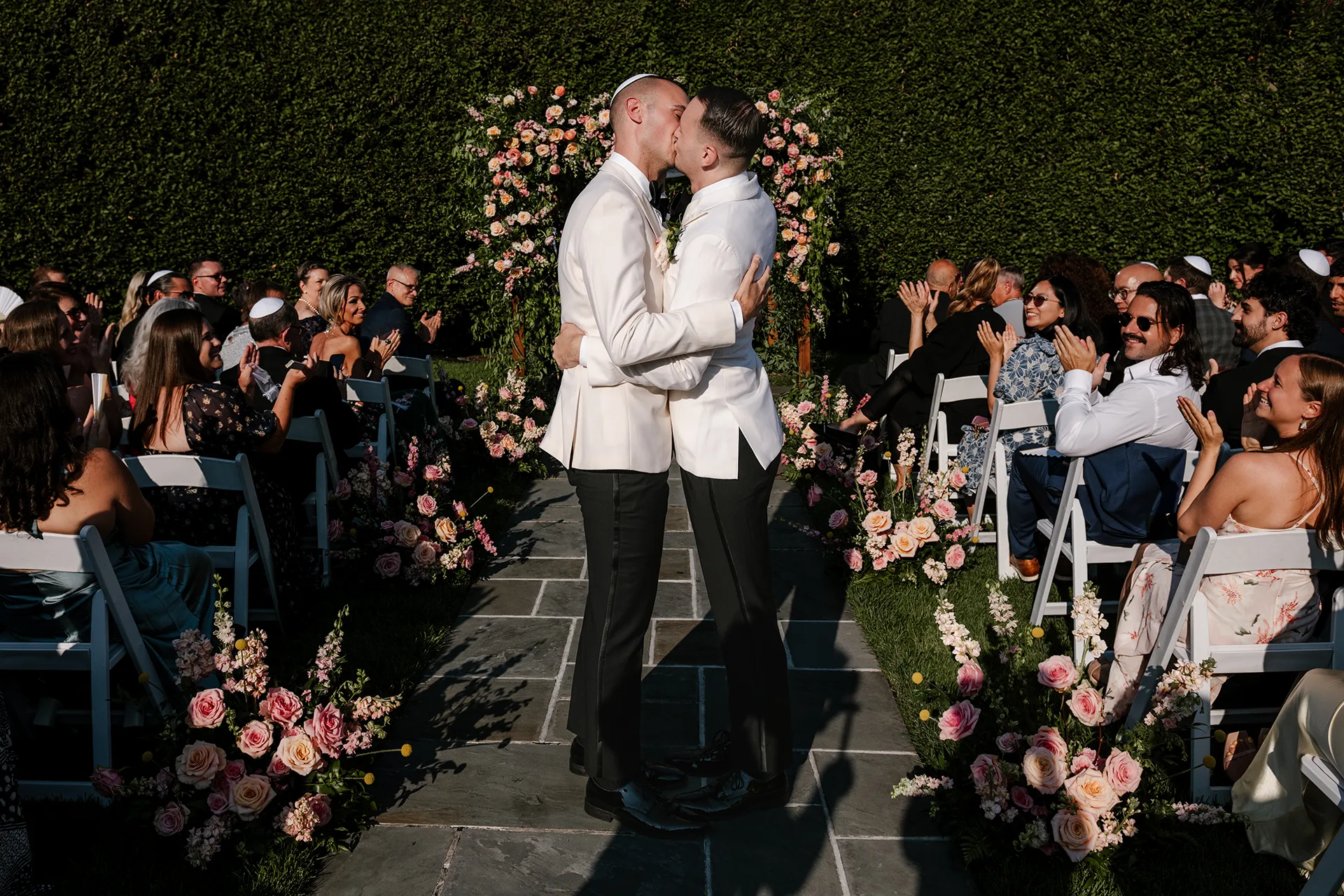Grooms kiss at outdoor wedding ceremony aisle framed by flowers and applauding guests.
