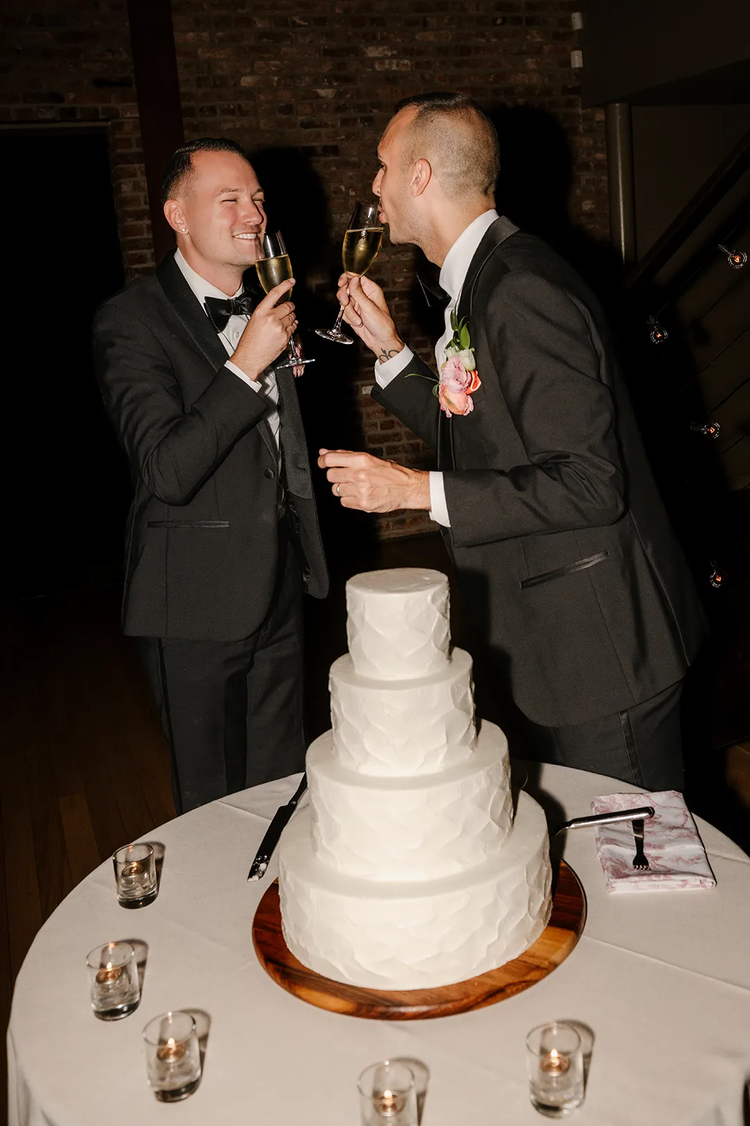 Two grooms toast champagne beside a three-tier white wedding cake.