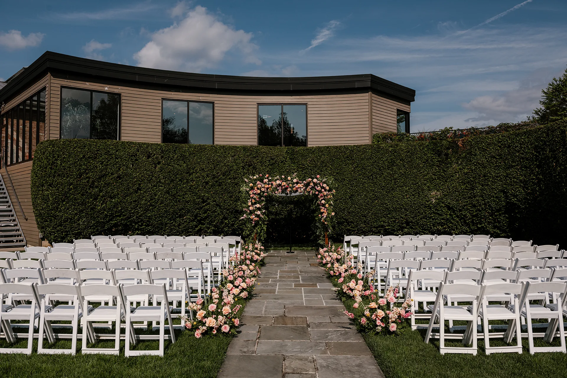 Outdoor wedding ceremony with white chairs, floral aisle, and flower-covered arch.