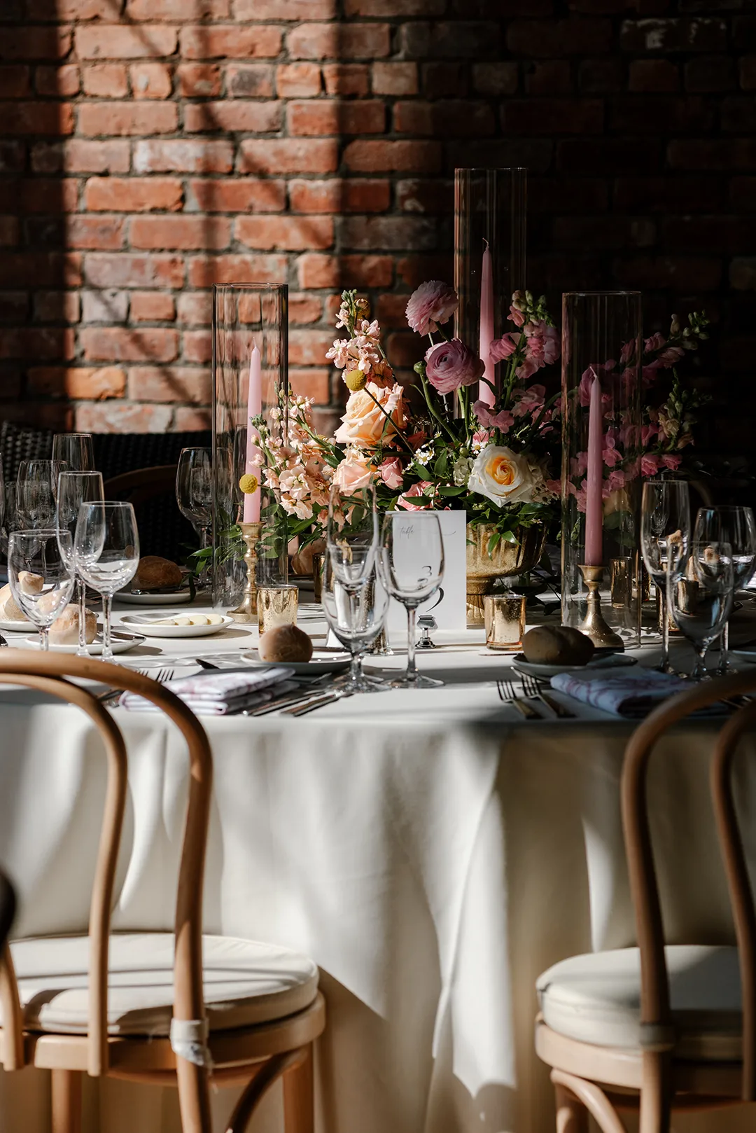 Elegant wedding table with floral centerpiece, candles, and place settings against a brick wall.