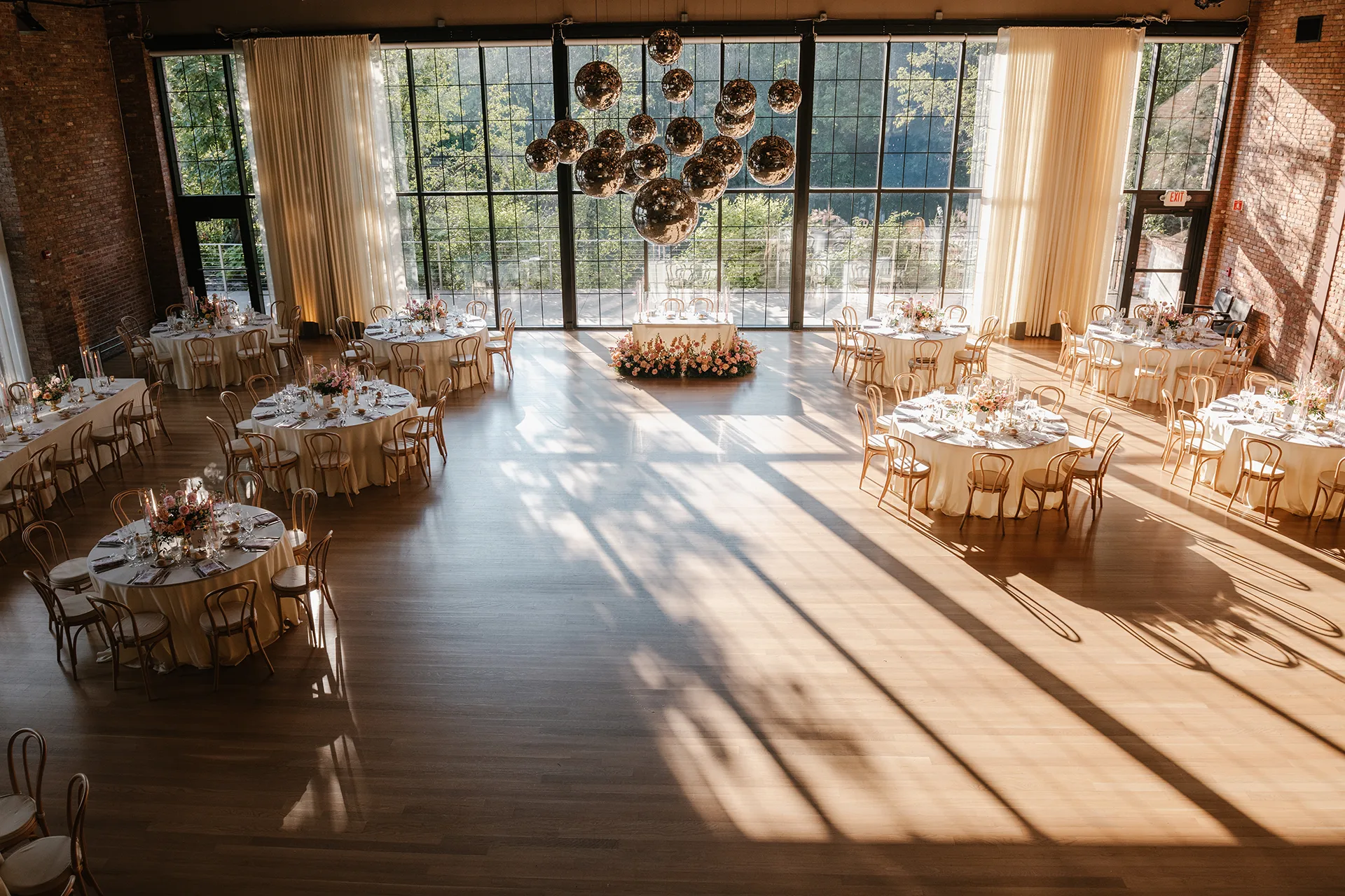Sunlit wedding reception hall with round tables, floral settings, tall windows, and hanging lights.