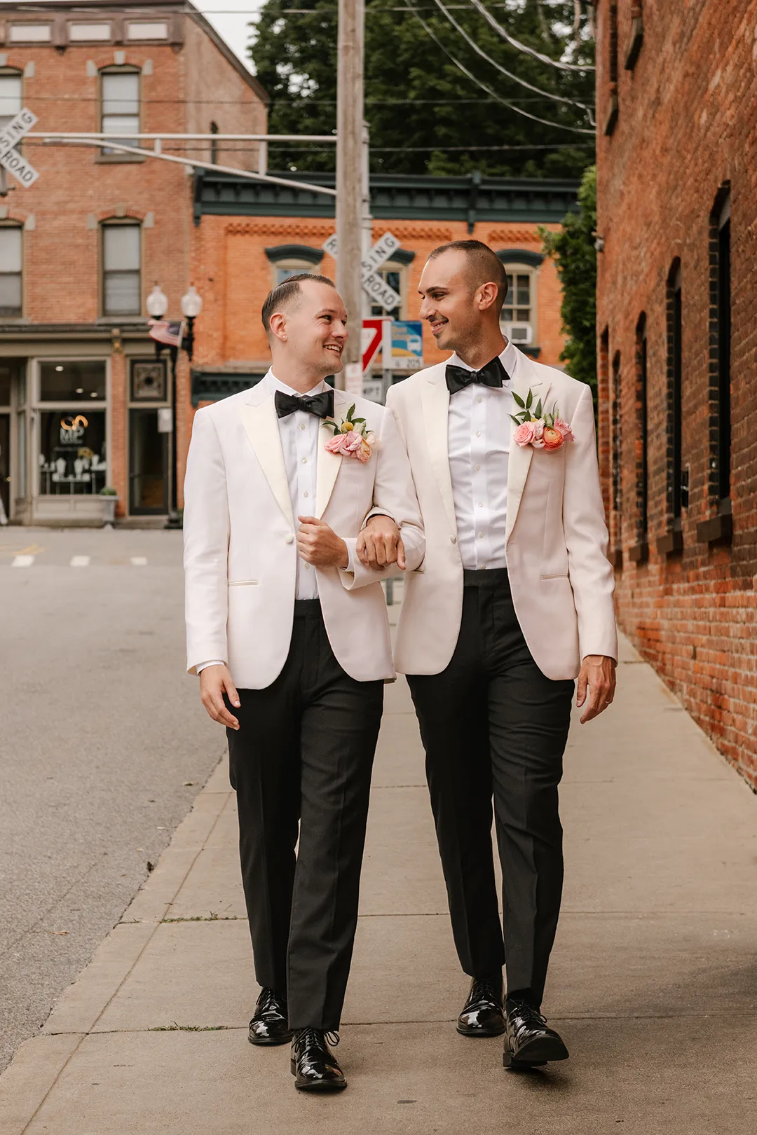 Two grooms in white jackets walk arm in arm down a city street, smiling at each other.