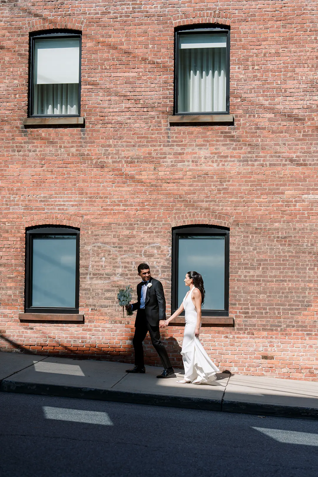 Bride and groom walk hand in hand past a brick building on their wedding day.
