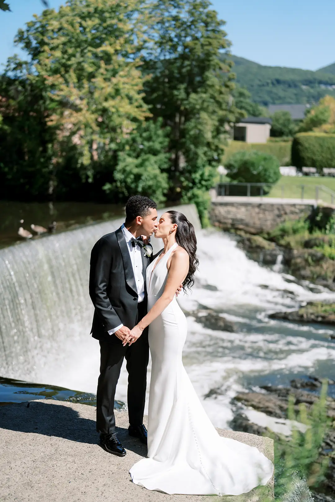 Newlywed couple kissing by a waterfall, wearing wedding attire with trees and hills behind.