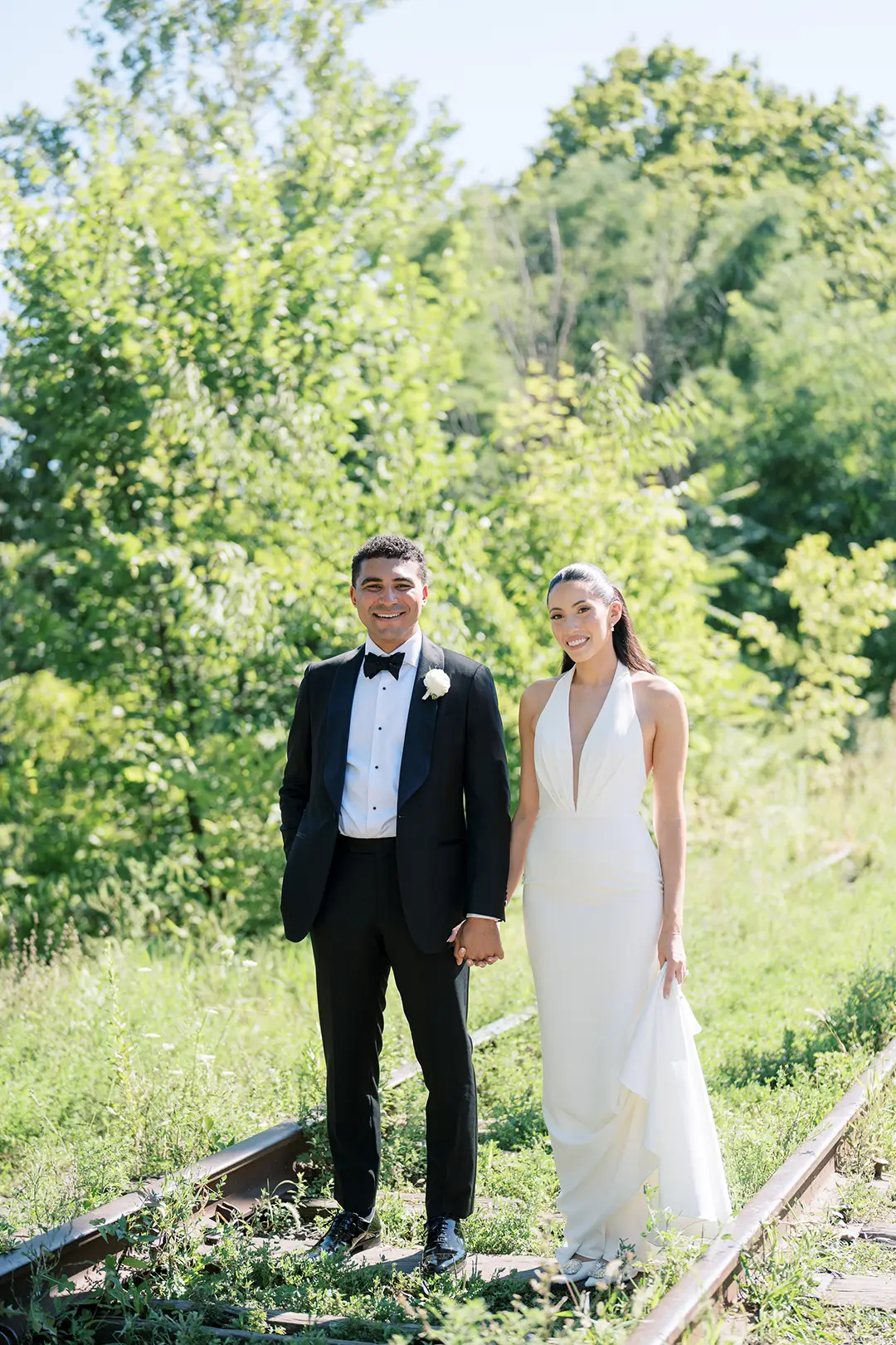 Wedding couple holding hands on sunlit path surrounded by lush green trees.