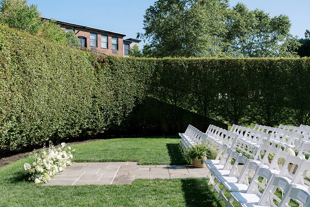 Outdoor garden ceremony with white chairs arranged on a lawn beside tall green hedges.