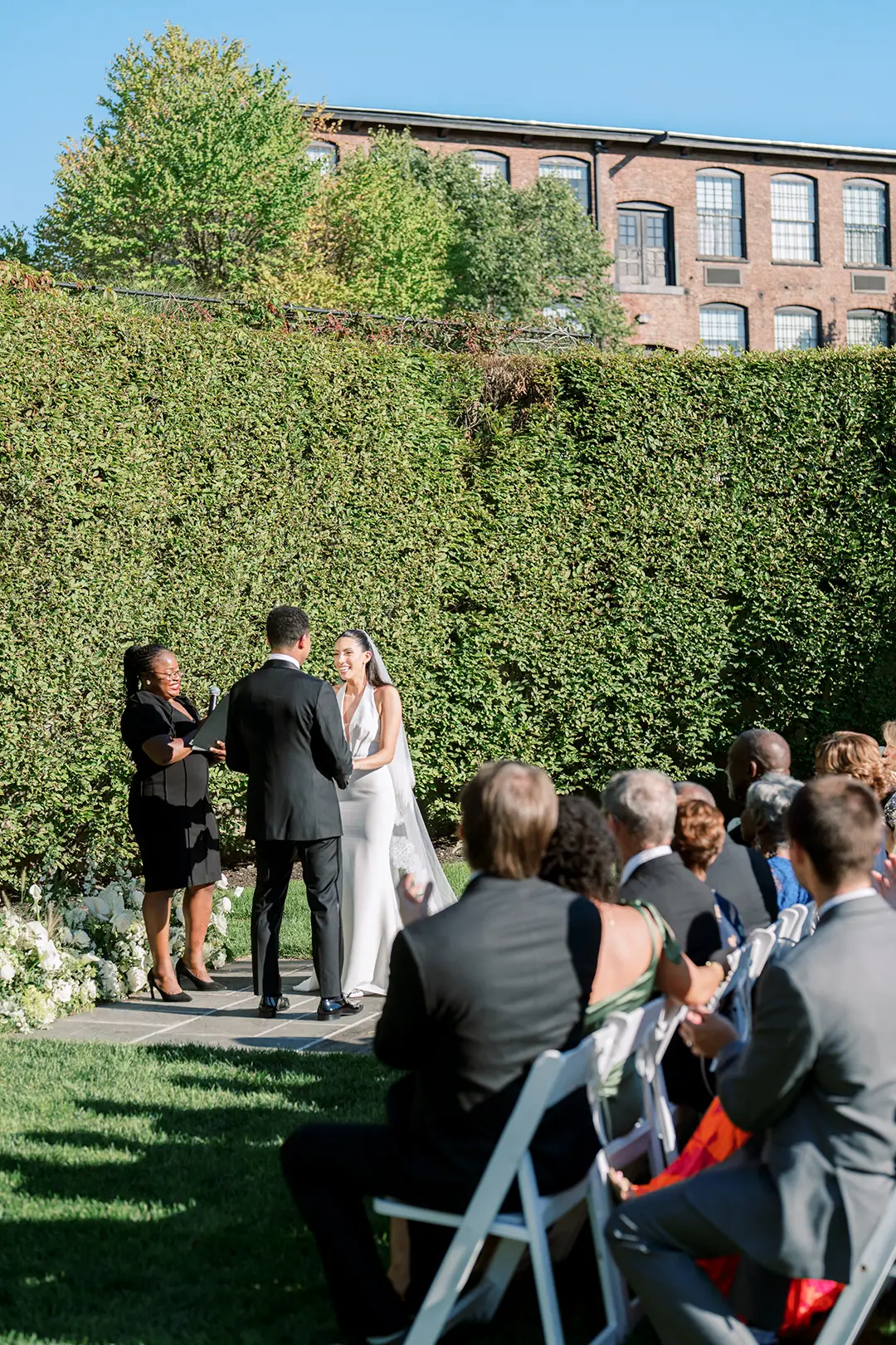 Outdoor wedding ceremony with couple exchanging vows before seated guests and greenery backdrop.