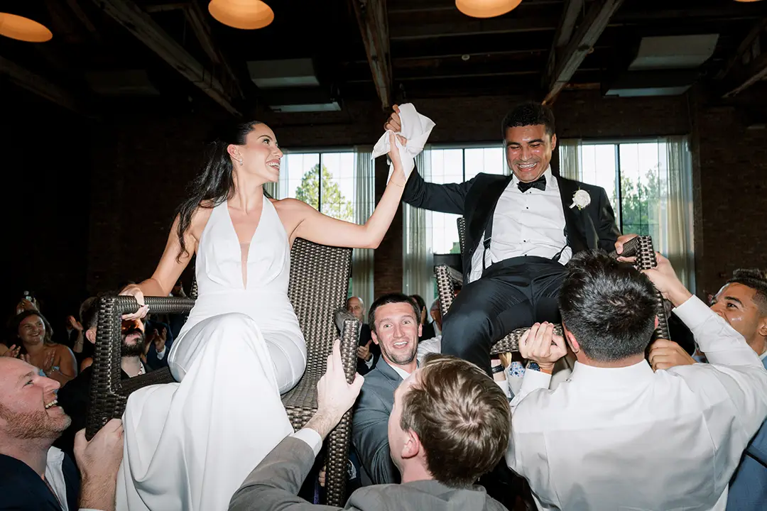 Guests lift bride and groom on chairs during a joyful wedding celebration indoors.