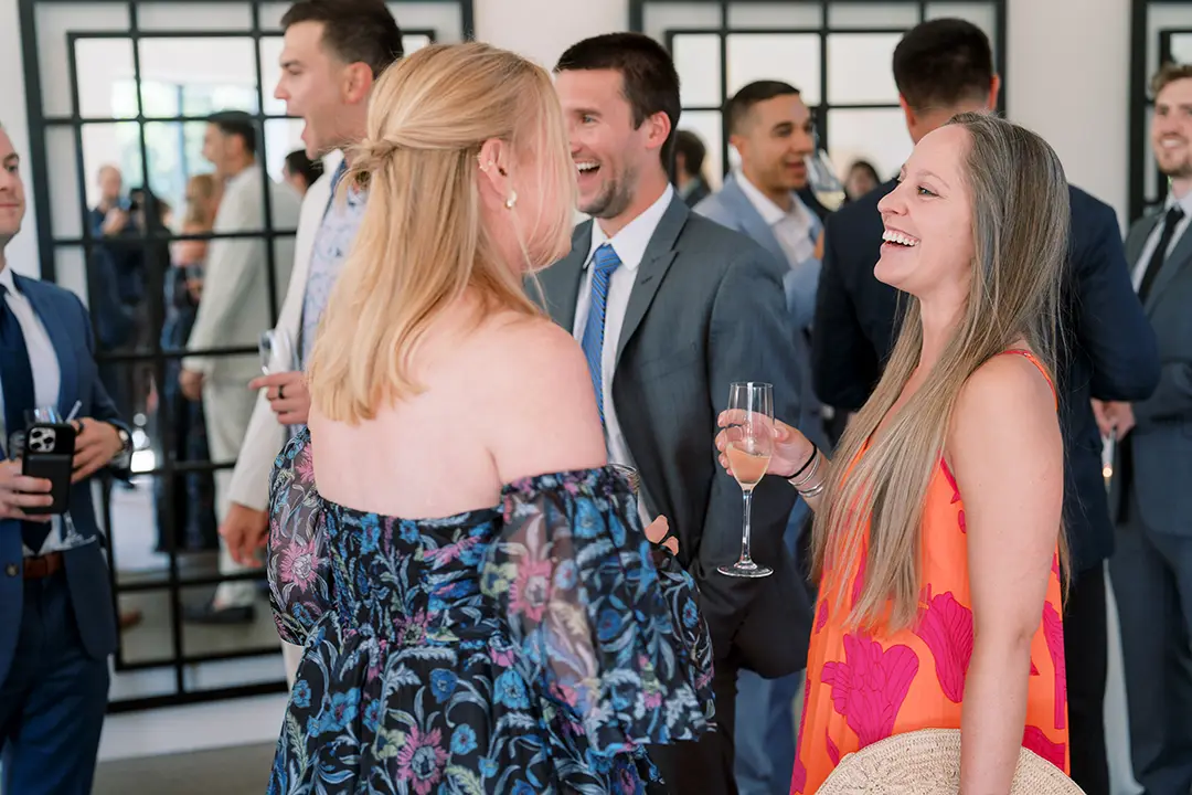 Guests laughing and chatting at a wedding reception, holding drinks in a bright indoor space.