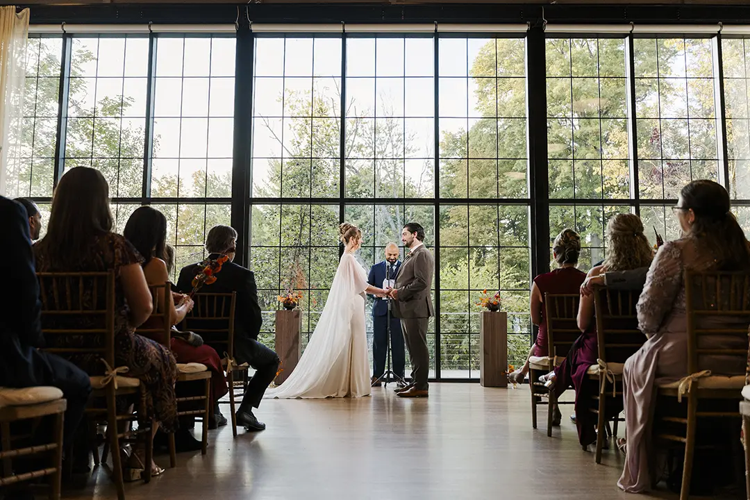 Couple exchanging vows indoors before seated guests, framed by tall windows and greenery outside.