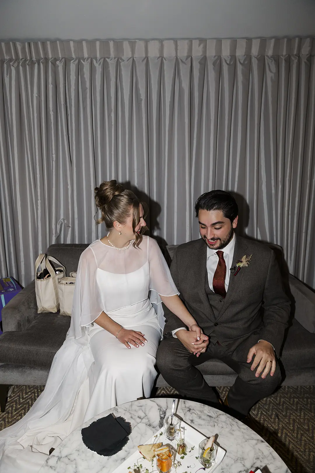 Bride and groom seated on a couch, holding hands and smiling indoors after their wedding.