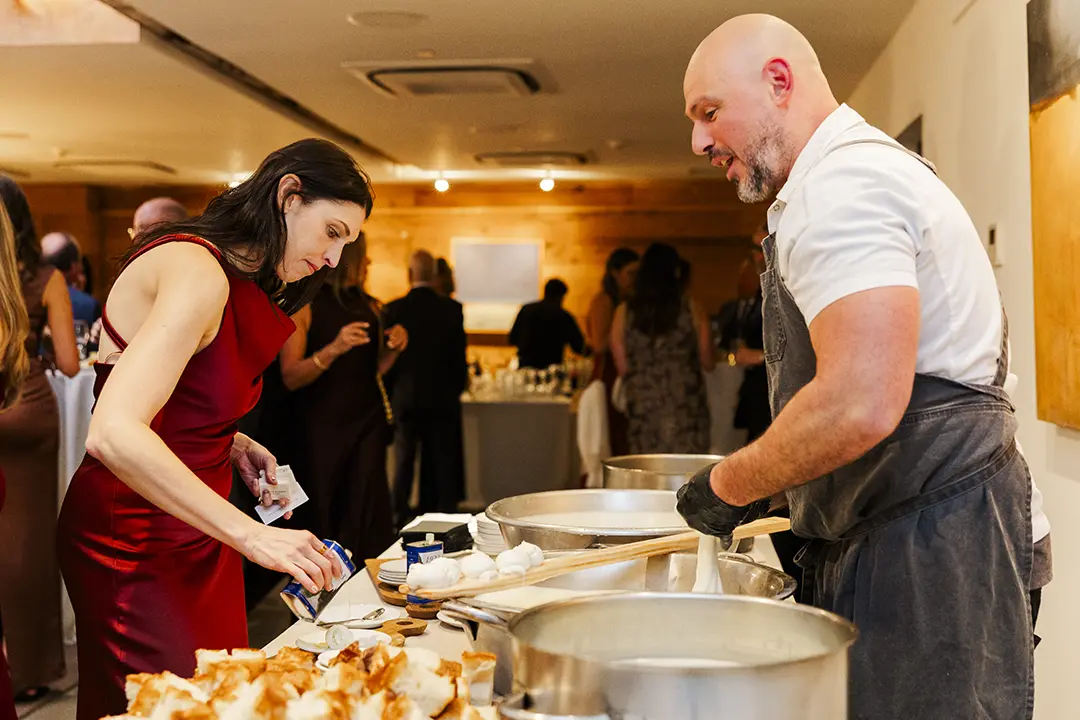 Chef serving food to a guest at an indoor event with guests mingling behind.