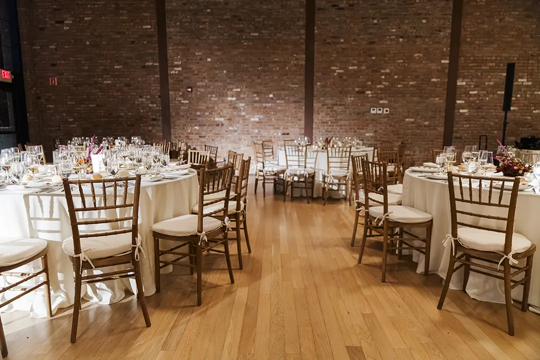 Elegant banquet room with round tables, white linens, wooden chairs, and exposed brick walls.
