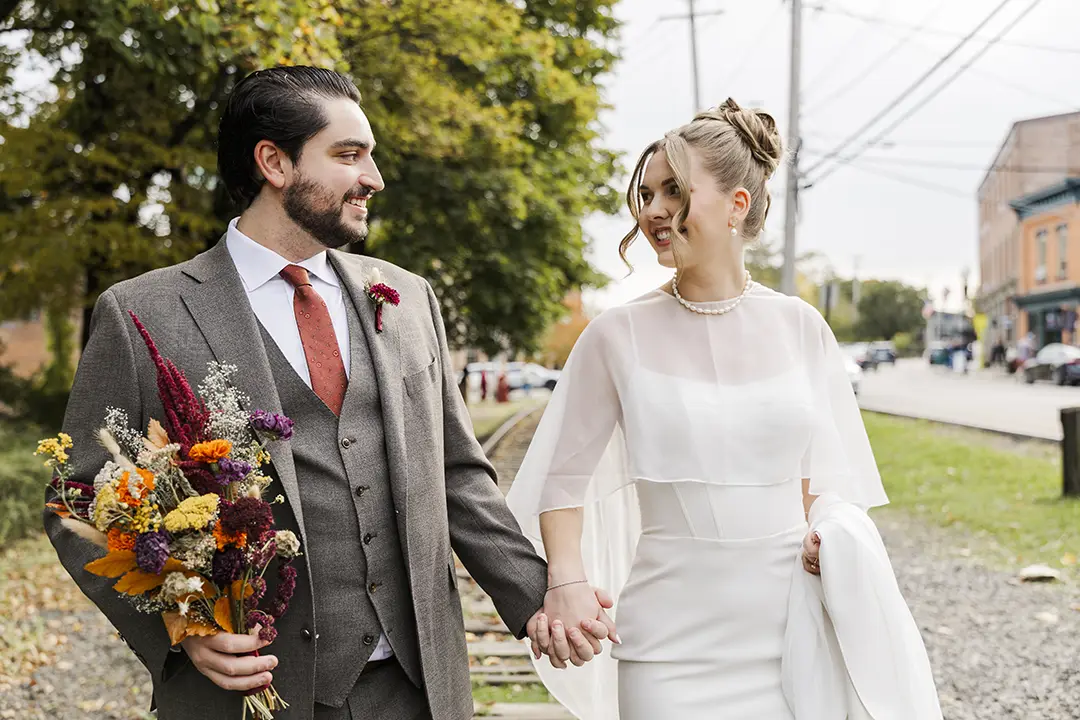 Bride and groom hold hands and smile while walking outdoors on their wedding day.