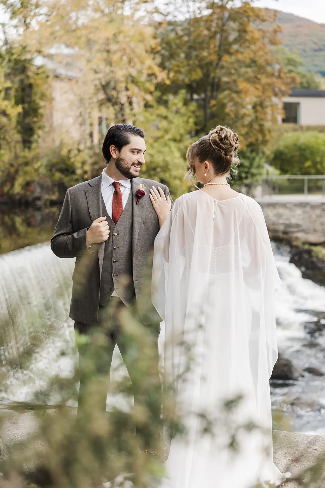 Wedding couple by a river, groom in suit smiling at bride in flowing white dress.
