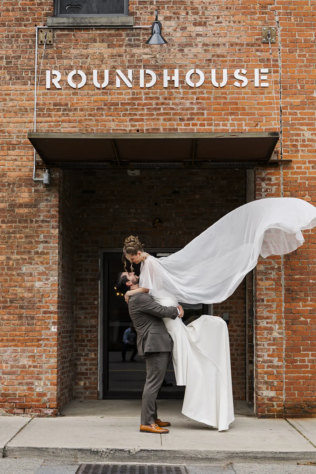 Groom lifts bride beneath roundhouse sign as her veil flows against a brick facade.