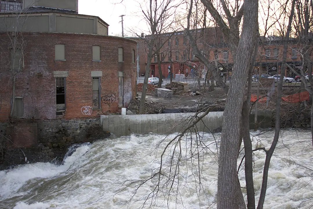 Old brick industrial building beside a fast-flowing river, with bare trees and construction nearby.