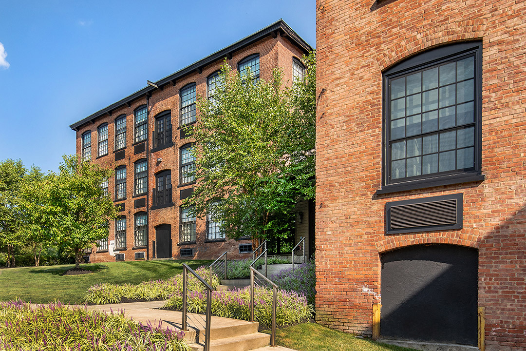 Historic brick mill buildings with large windows, trees, and landscaped walkway on a sunny day.