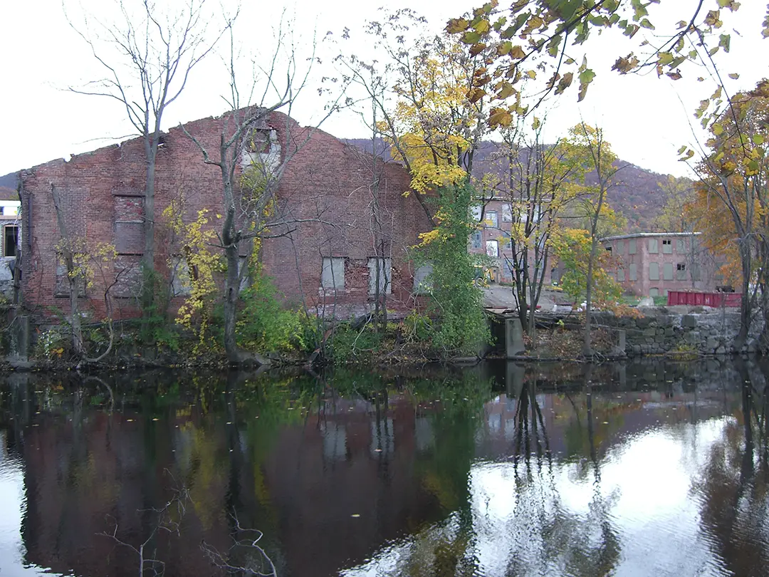Abandoned brick mill by a calm river, reflected water, autumn trees, and hillside backdrop.
