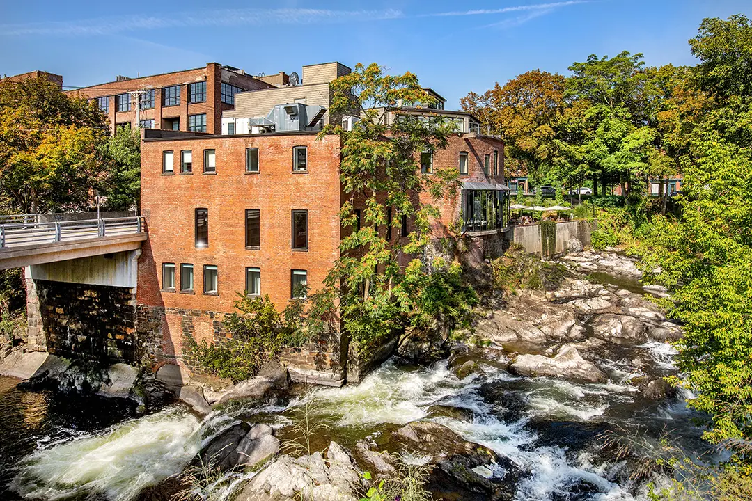 Brick mill building over a rushing river, surrounded by trees under a clear blue sky.