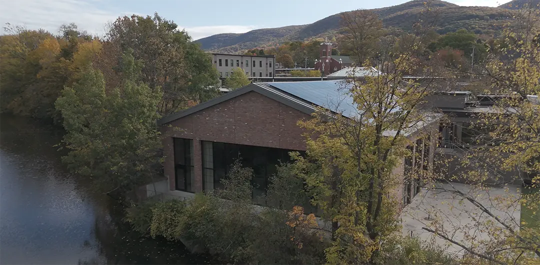 Brick building beside a river with trees and mountains in the background during autumn.