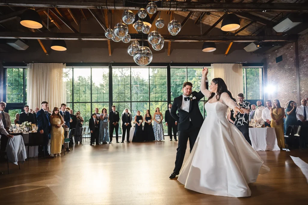 A couple enjoys their first dance in the waterfall room at the roundhouse surrounded by applauding friends and family under disco balls, exposed wooden beams, with floor-to-ceiling windows showing hudson valley nature.